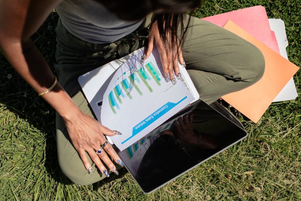 Woman working outdoors on grass with a laptop and documents, reviewing sales data.